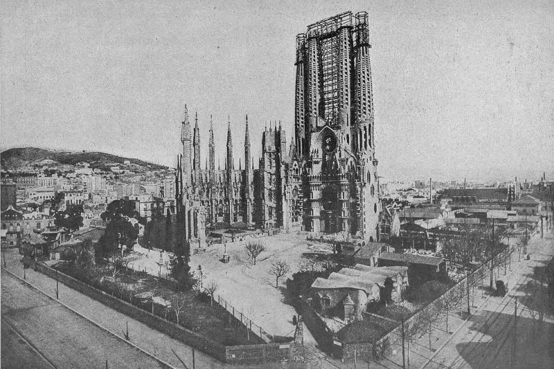 Photographie en noir et blanc du chantier de la Sagrada Familia en 1927. C'est une vue d'ensemble du terrain, avec quelques arbres, des petites constructions, la seule façade terminée avec des échafaudages et la ville en fond.