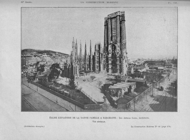 planche issue de la revue la construction moderne montrant une photographie en noir et blanc du chantier de la Sagrada Familia. On voit le terrain avec quelques arbres, des petites constructions et la seule façade déjà construite, encore soutenue par des échafaudages au niveau des tours, et au dernier plan on voit la ville.